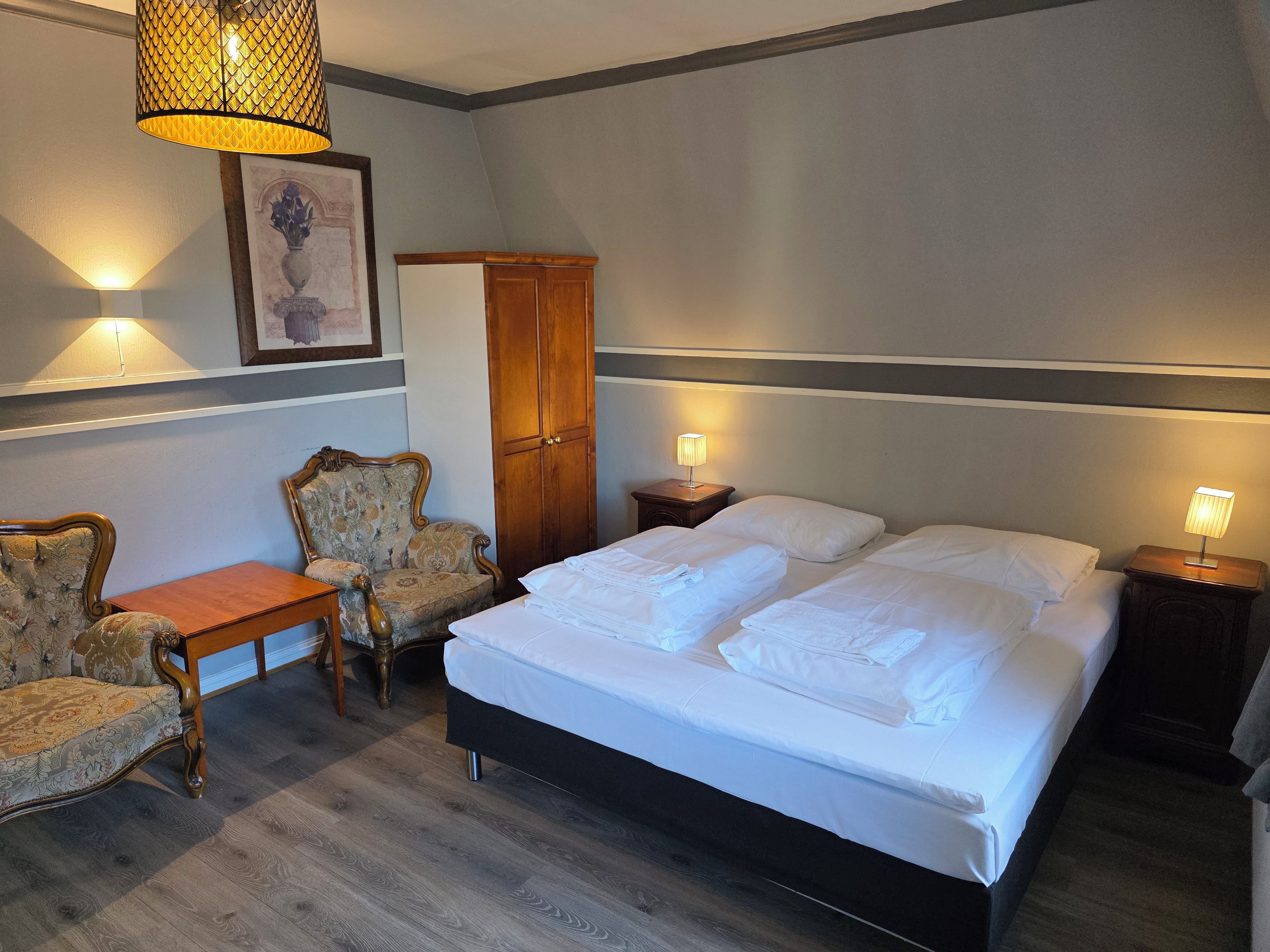Une chambre d’hôtel avec un grand lit, deux tables de chevet, une armoire en bois, une chaise et une lampe.