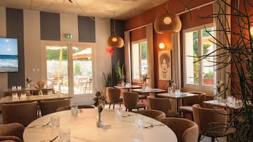 Un restaurant avec des tables rondes, des chaises recouvertes de tissu marron, des lampes en osier suspendues au plafond et une vue sur l’extérieur grâce à de grandes fenêtres.