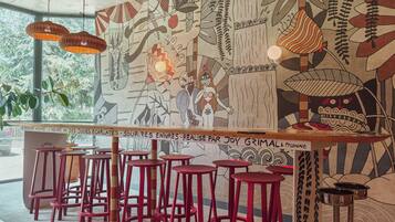 Een café-interieur met rode barkrukken, een muurschildering met abstracte kunst en hangende rieten lampen.