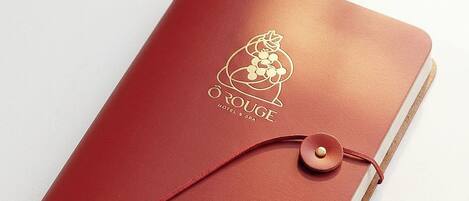 A red leather-bound notebook with a cork band and a button closure, featuring a logo and text on the cover.