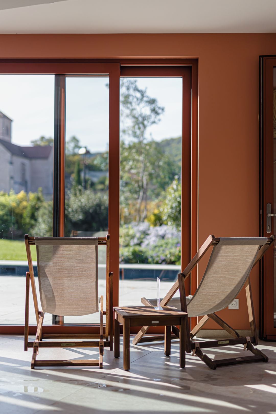 A room with two wooden chairs and a small table, a sliding glass door leading to an outdoor area, and a view of greenery and a building outside.