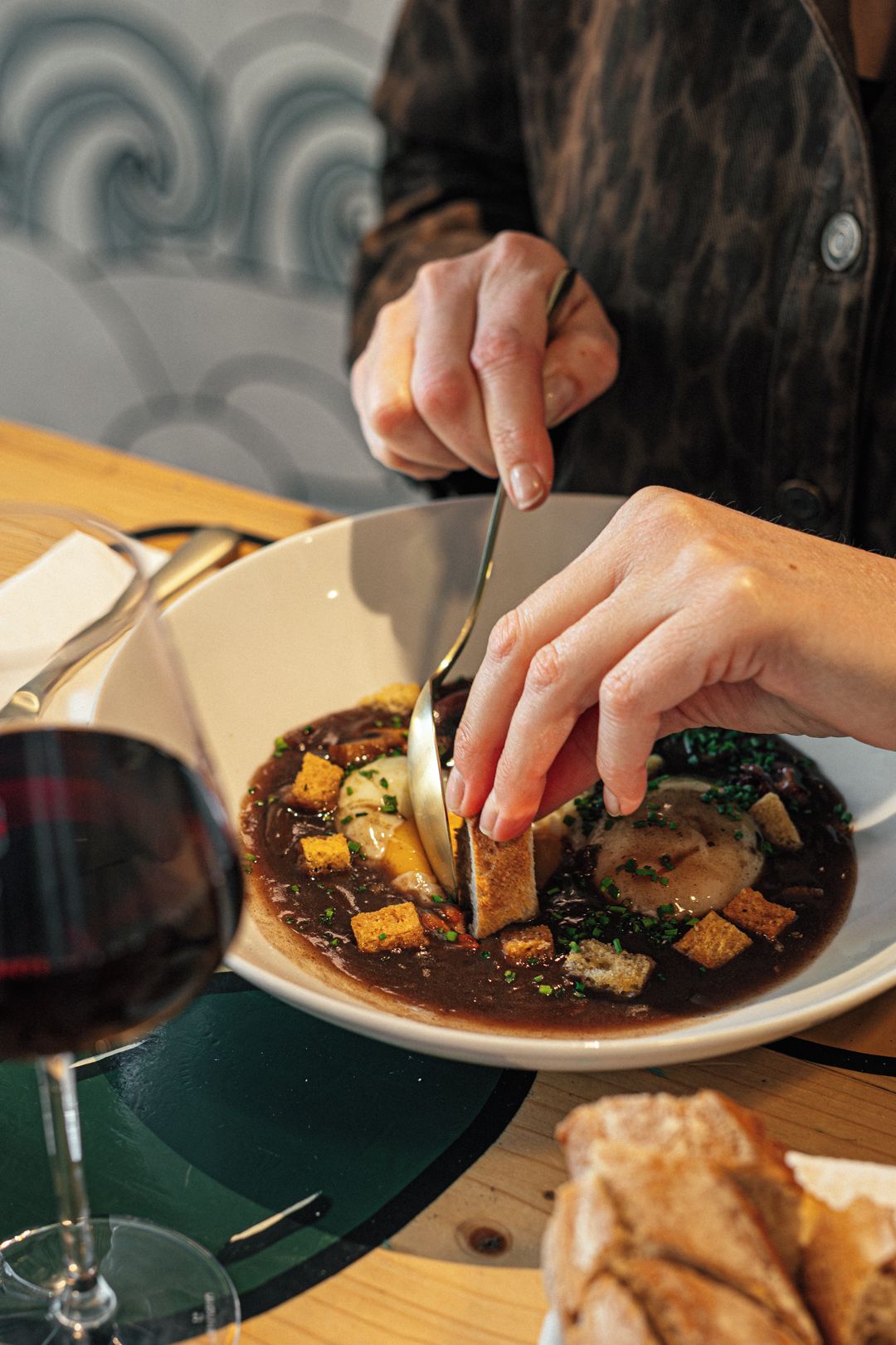 Une personne qui coupe de la nourriture avec un couteau et une fourchette sur une assiette, avec un verre de vin rouge et du pain sur la table.