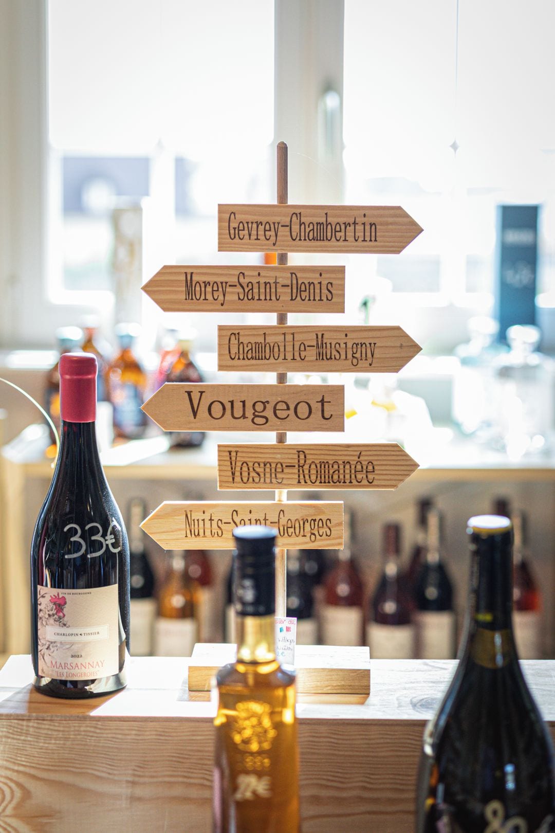 Ein hölzernes Wegweiser mit Namen von Weinregionen, die in verschiedene Richtungen zeigen, eine Flasche Marsannay Wein und weitere Weinflaschen im Hintergrund.