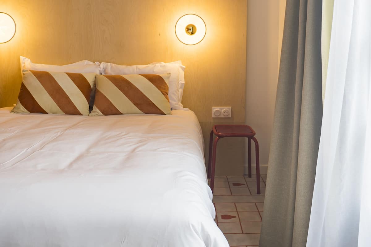 A hotel room with a bed, a small stool, a wall-mounted light, and a curtain.