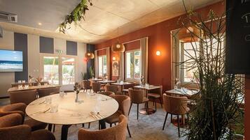 A restaurant with round tables, chairs, hanging lights, and a large TV screen.