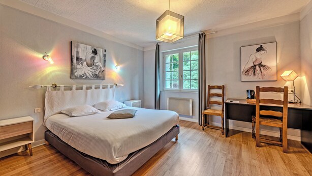 A bedroom with a bed, a desk, and chairs. There are paintings on the walls and a window with curtains. - Hotel De la Corniche (Brest)