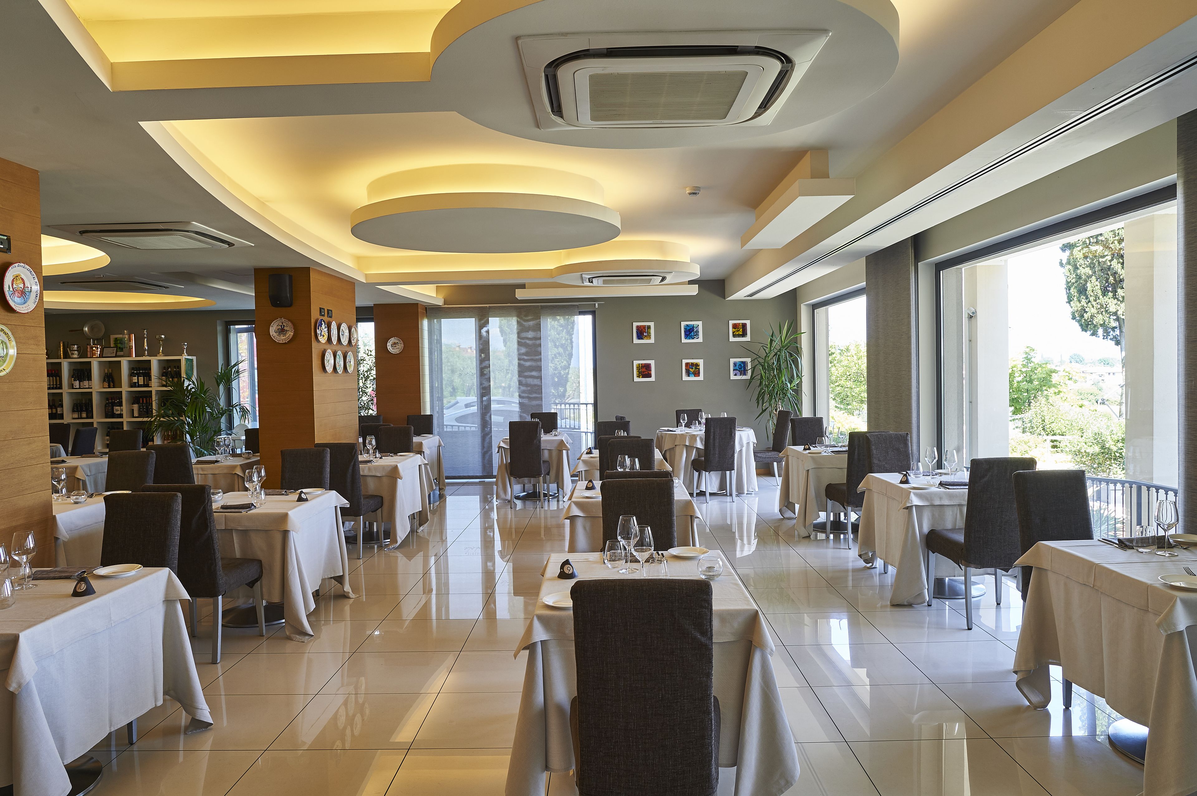 Ein Restaurant mit mehreren Tischen und Stühlen, einer Decke mit eingelassener Beleuchtung und großen Fenstern mit Blick auf Grünflächen.