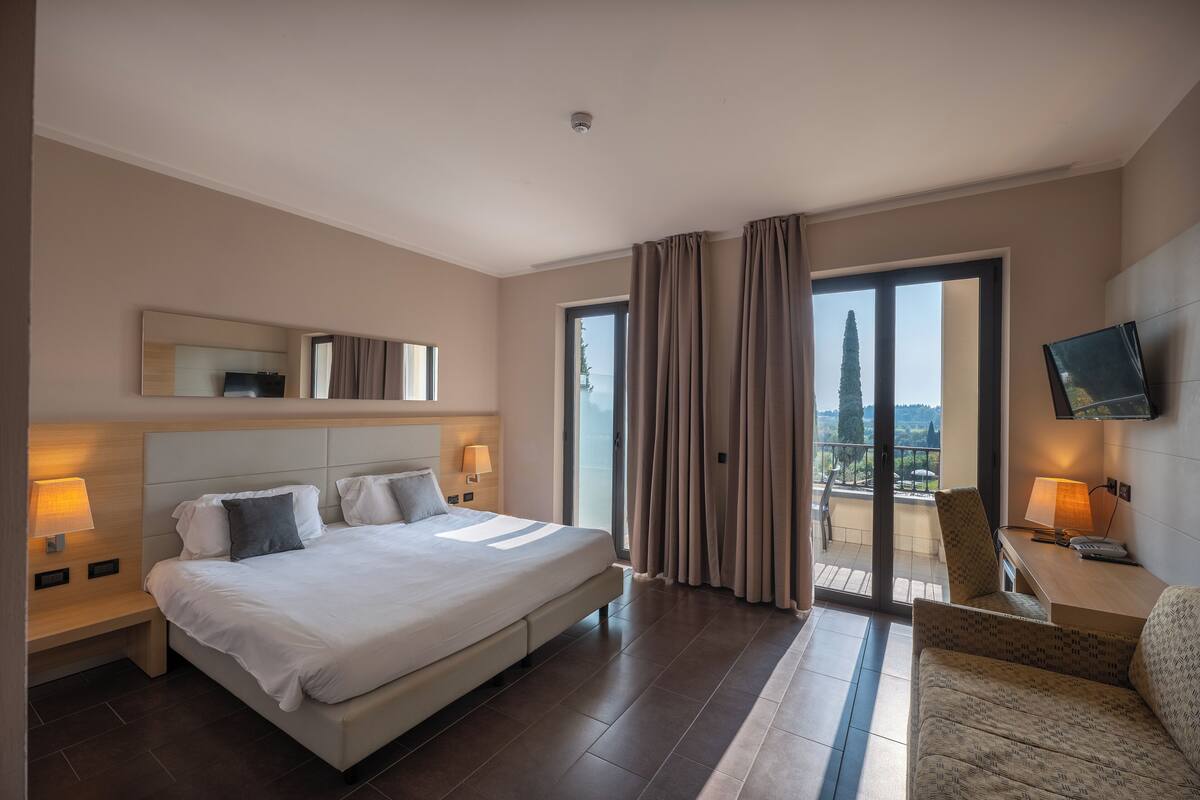 Una habitación de hotel moderna con una cama grande, un escritorio con silla, televisión y un balcón con cortinas.