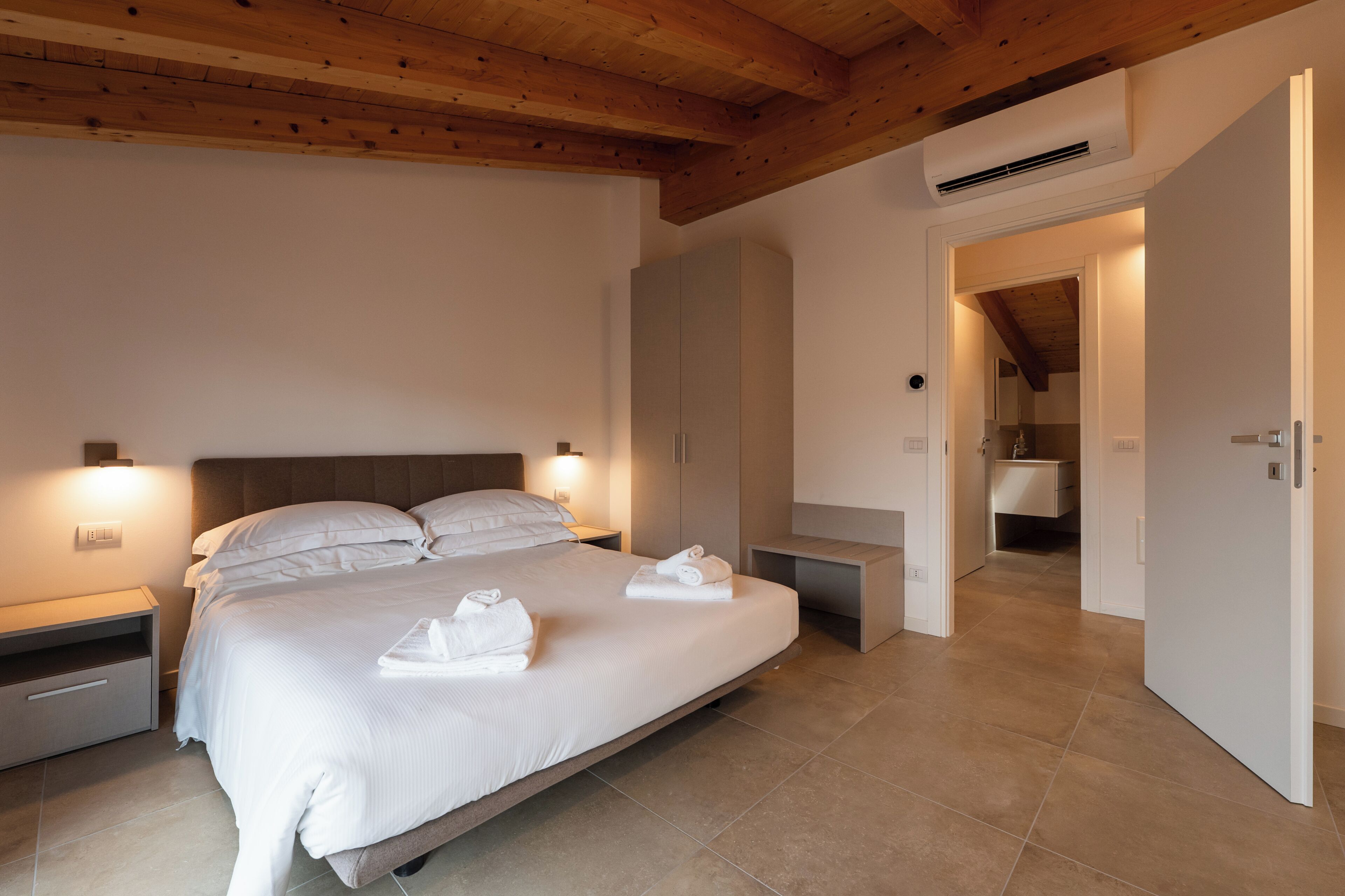 Une chambre à coucher avec un grand lit, des tables de chevet, une armoire et une salle de bain visible à travers une porte ouverte.