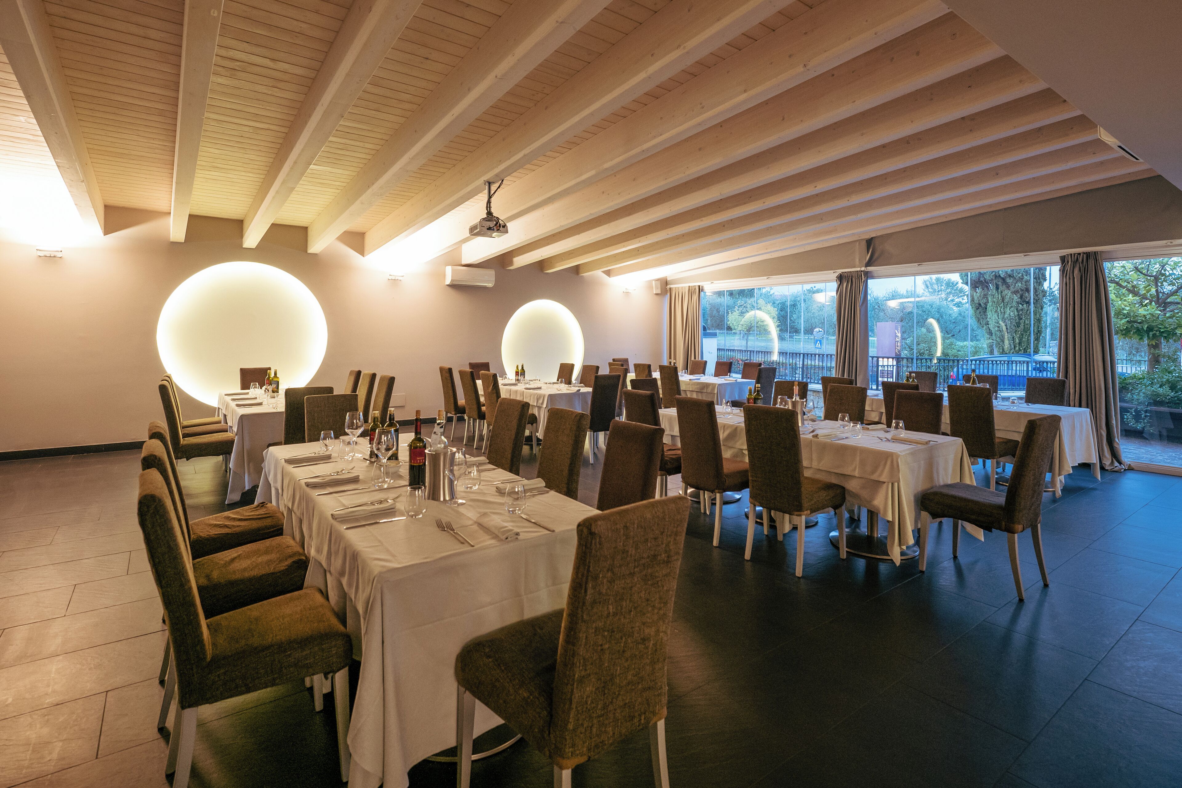 A dining area with multiple tables and chairs, large round light fixtures on the ceiling, and a view through large windows.