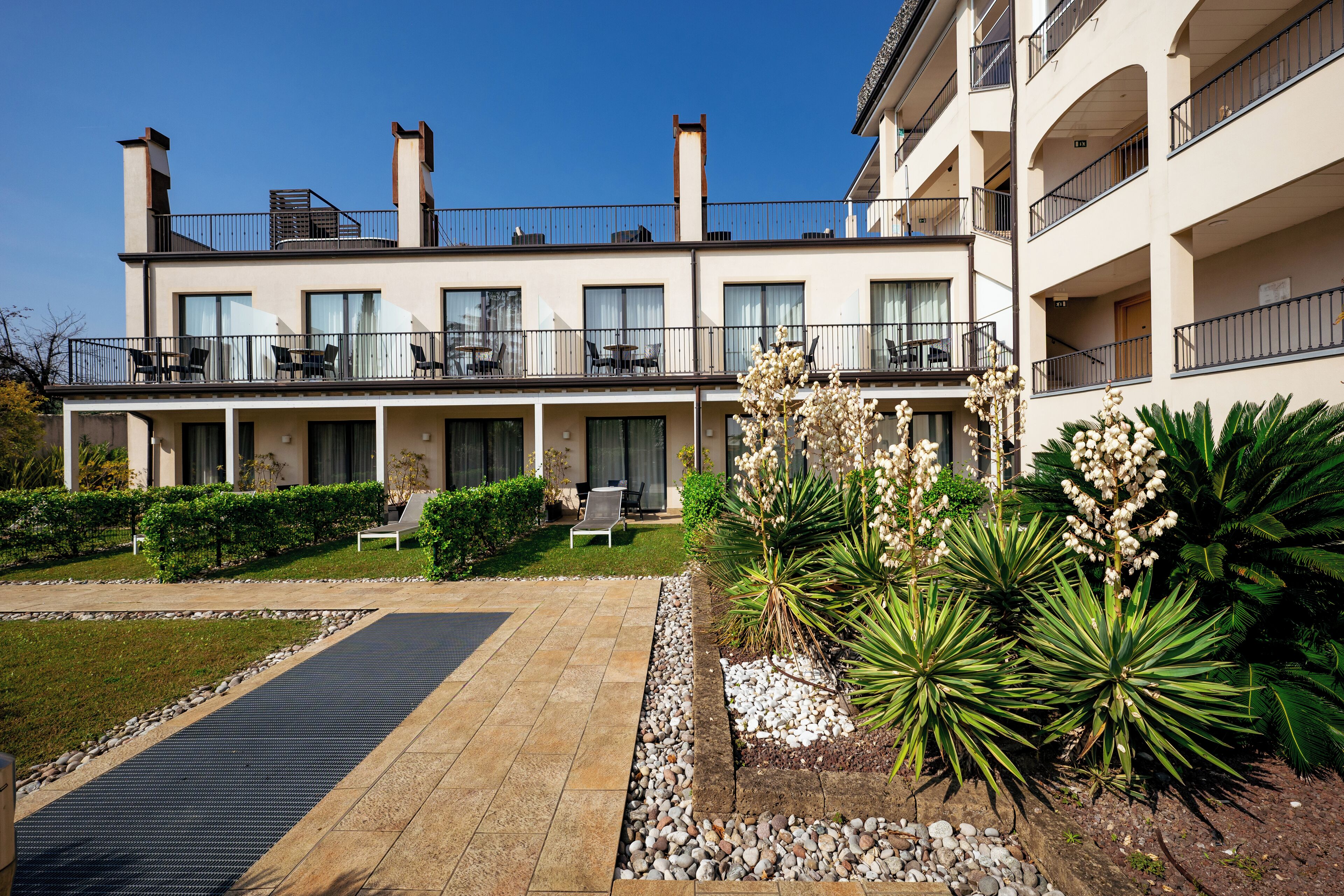 A modern hotel with a covered outdoor seating area, a swimming pool, and landscaped gardens.