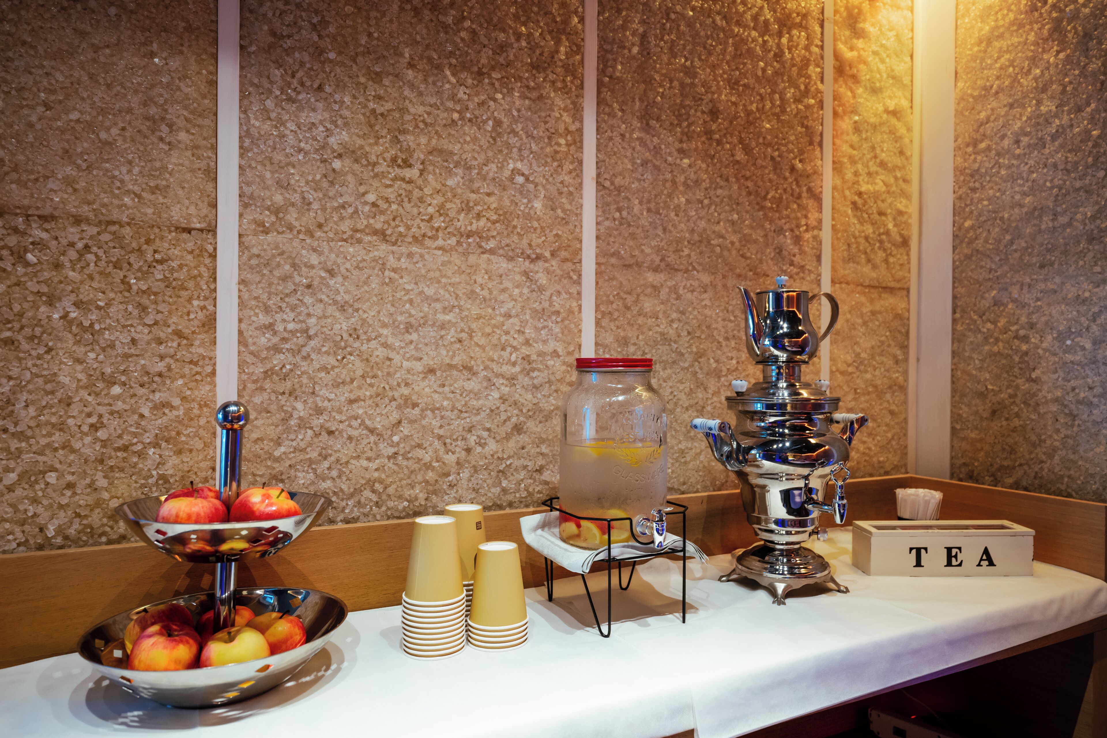 A buffet setup with a fruit bowl, stacked cups, a beverage dispenser, and a tea station.