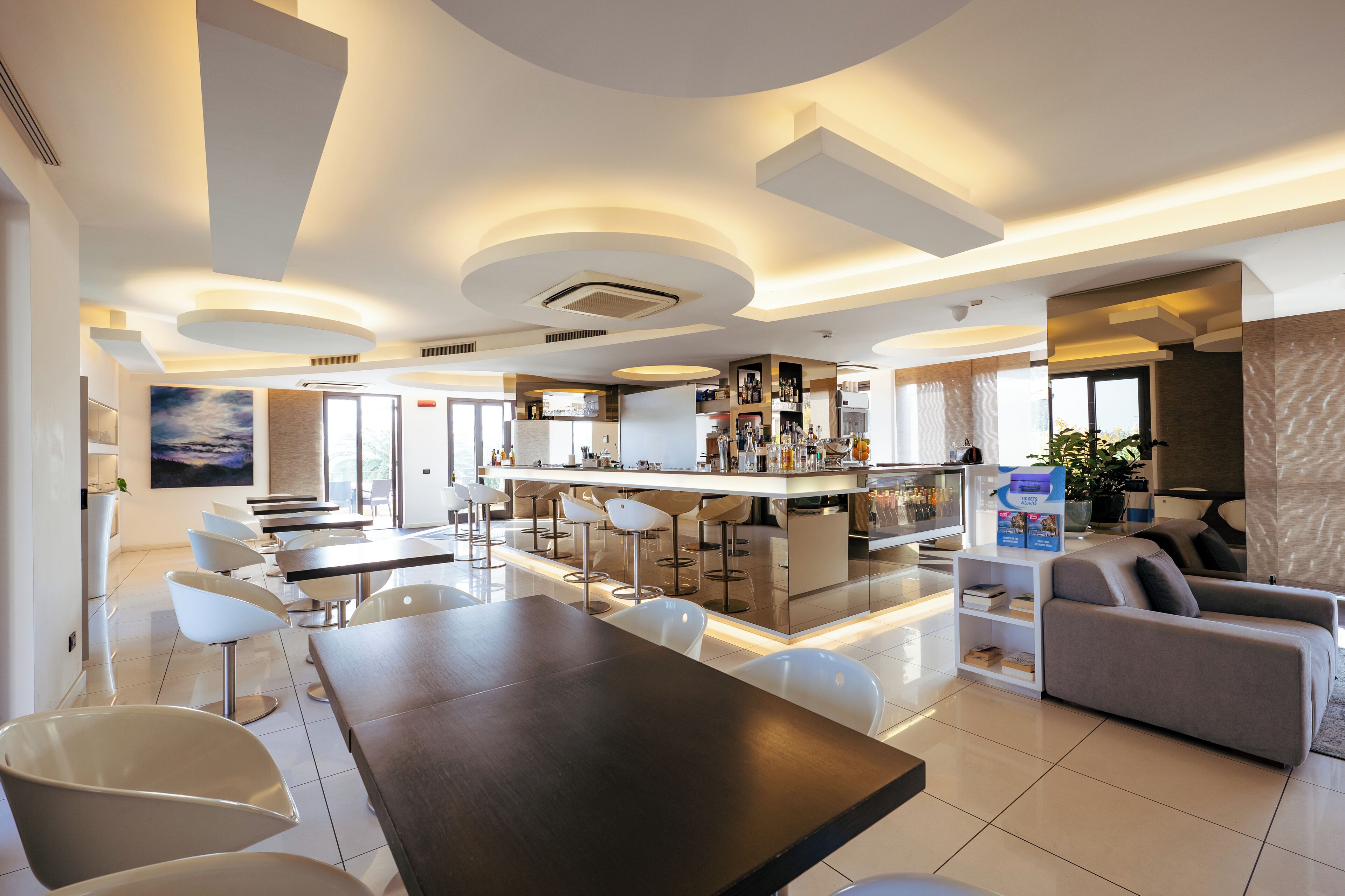 Un moderno hall de hotel con un bar central, zona de estar y un gran ventanal que proporciona luz natural.