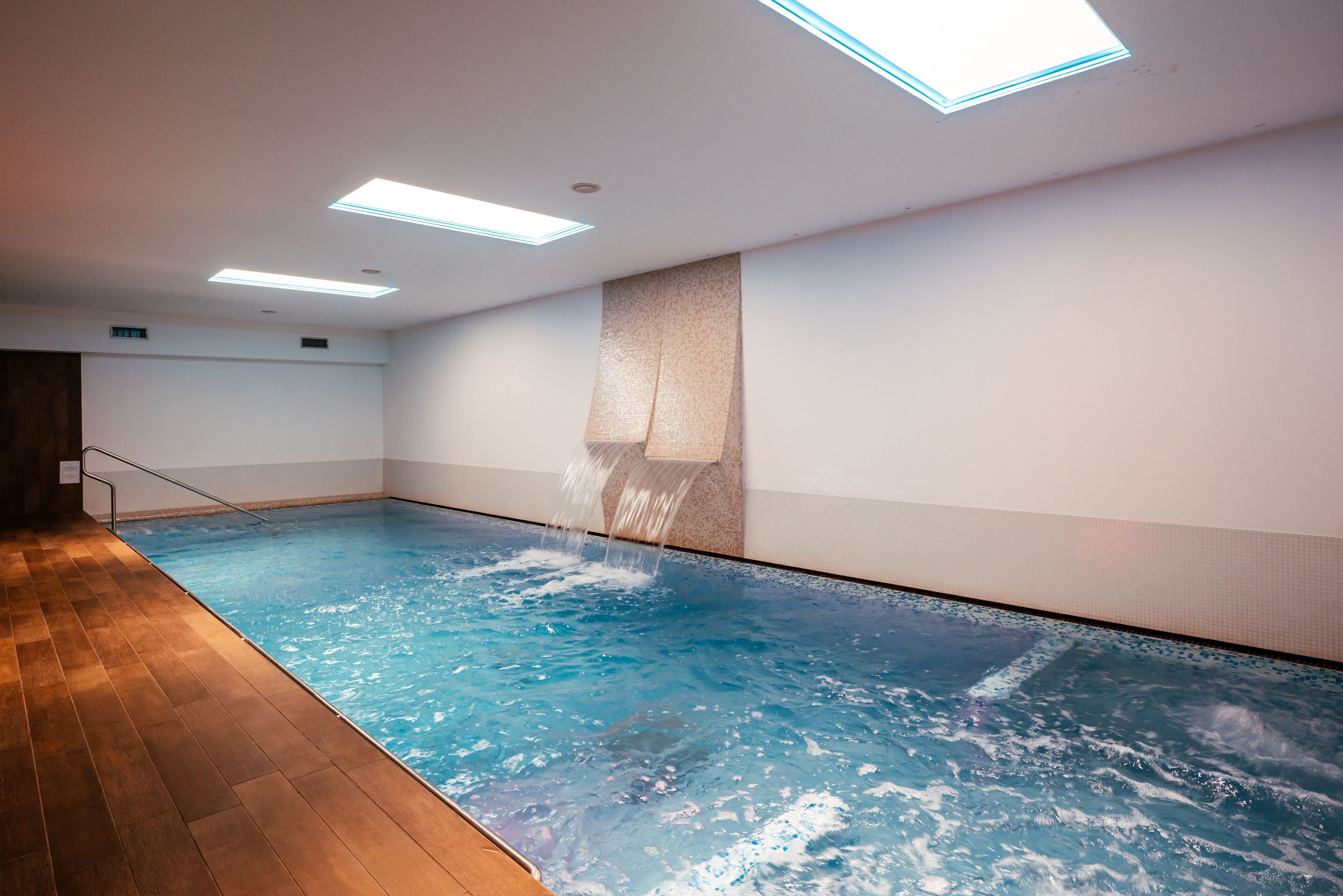 An indoor swimming pool with a wooden floor, a water feature, and recessed lighting.