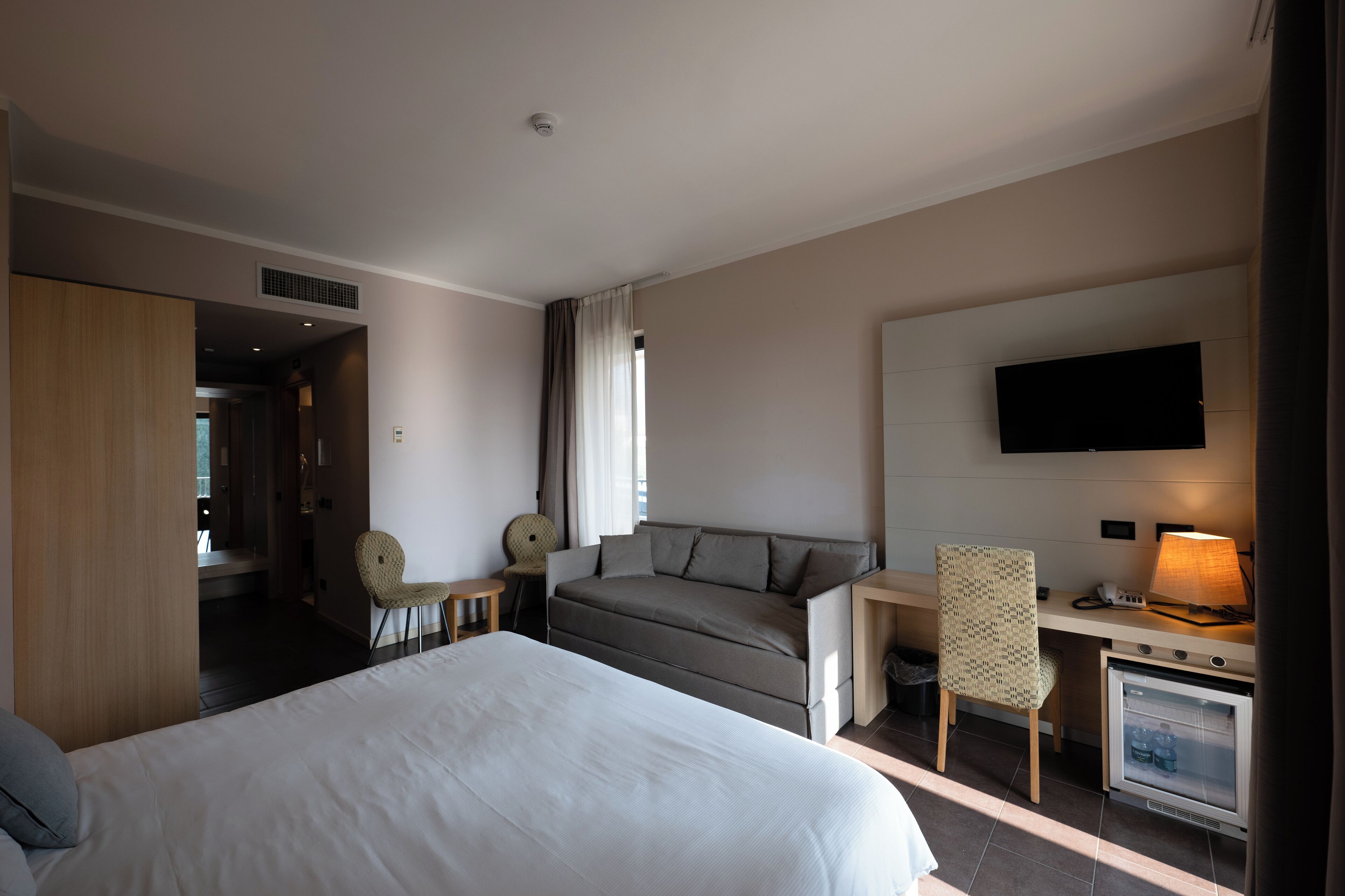 Ein Hotelzimmer mit einem großen Bett, einer Sitzecke, einem Schreibtisch mit Lampe, einem Fernseher und einem kleinen Kühlschrank.