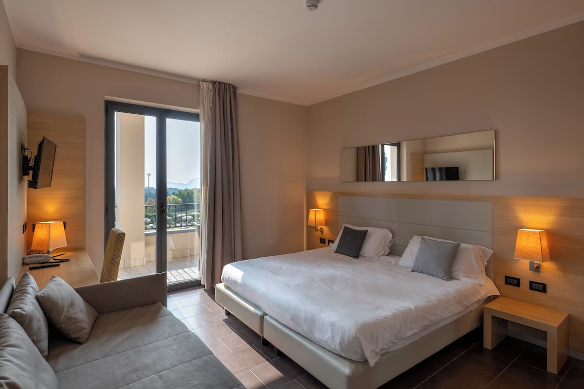 Una habitación de hotel moderna con una cama grande, un sofá, un balcón con cortinas y un televisor instalado en la pared.