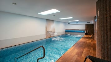 An indoor swimming pool with clear water, wooden flooring, and a bench along the wall.
