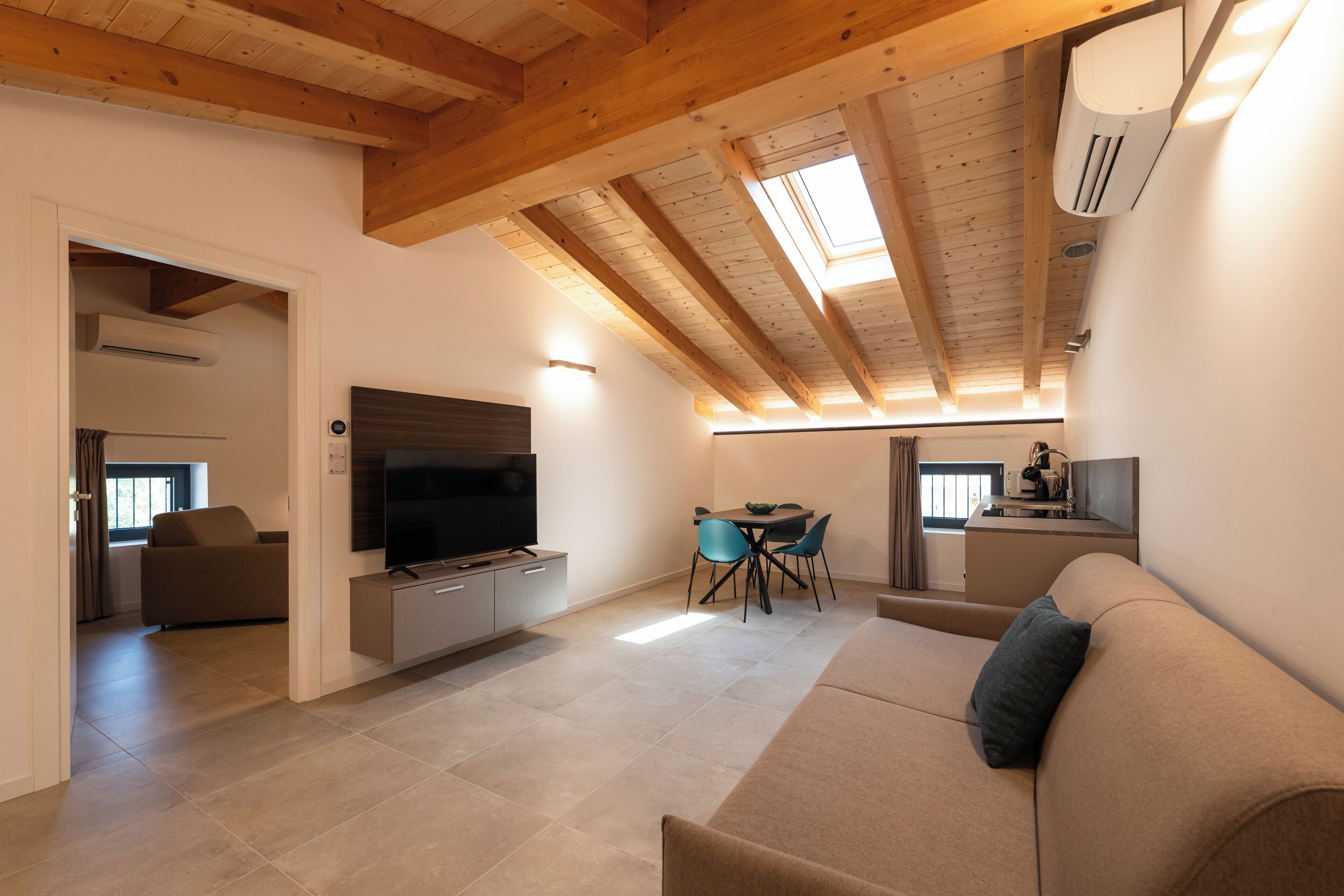 A modern living room with a flat-screen TV, a sofa, a dining area with chairs, and a kitchen area with a sink and a kettle.