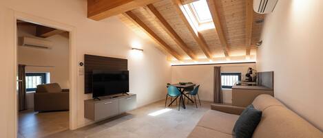 A modern living room with a flat-screen TV, a sofa, a dining area with chairs, and a kitchen area with a sink and a kettle.
