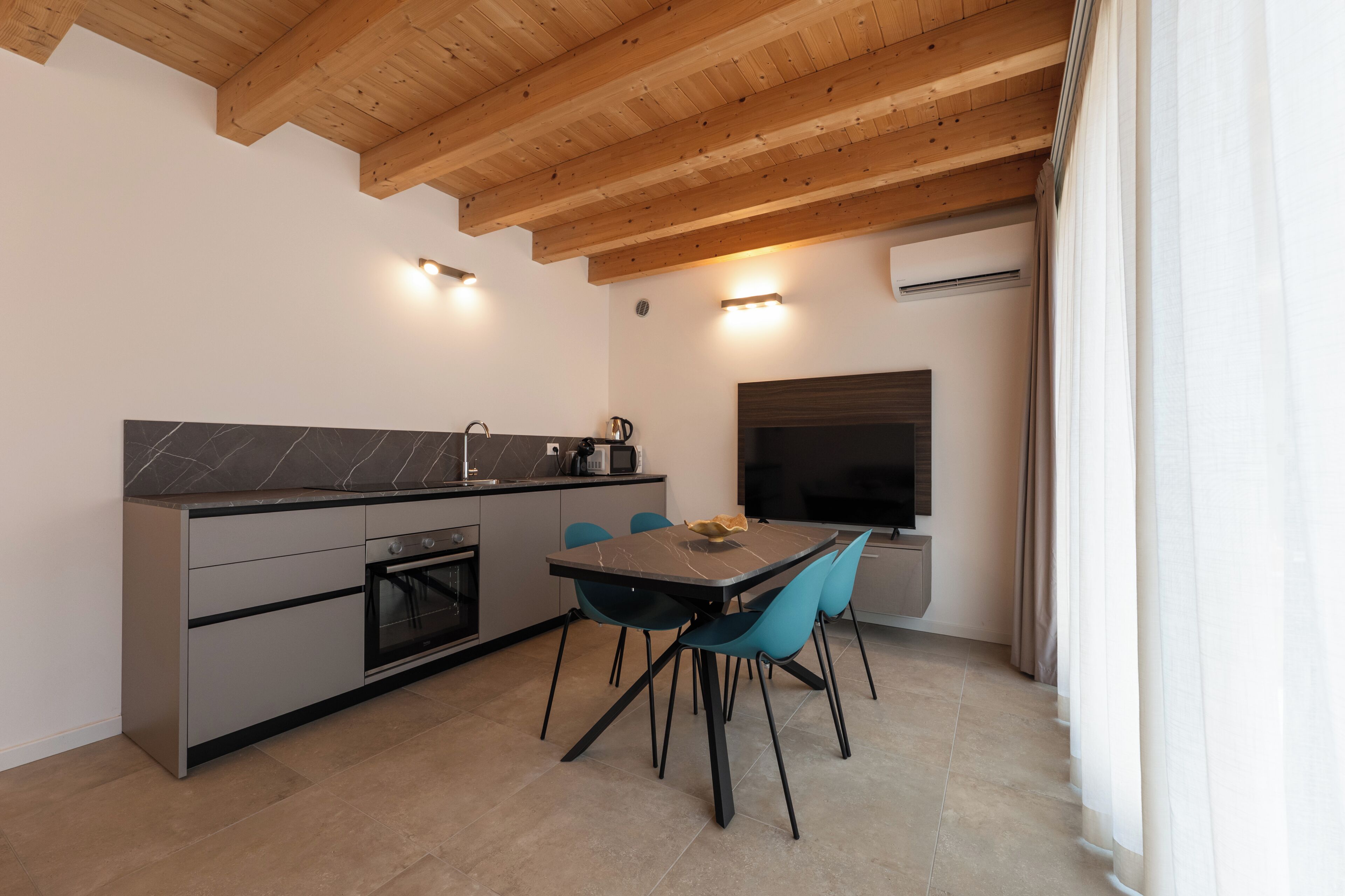 A modern kitchen with a built-in oven, a microwave, and a coffee maker. There is a dining table with chairs and a flat-screen TV mounted on the wall.