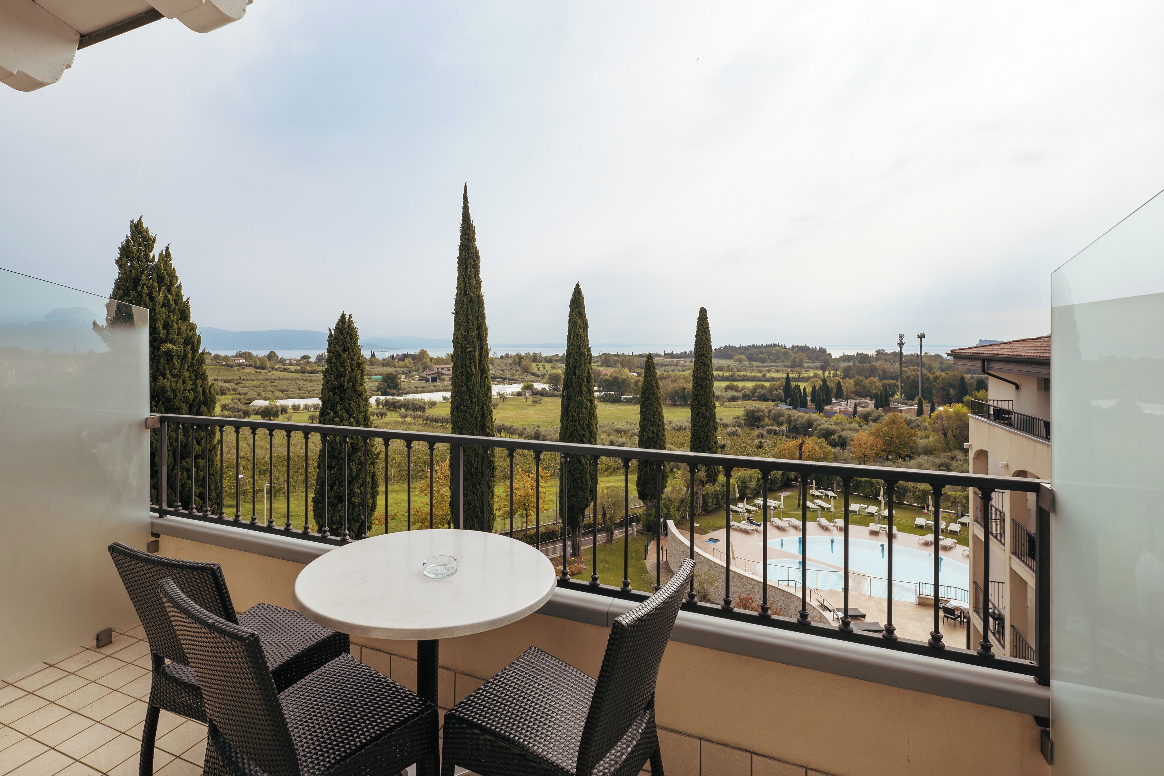 A balcony with a round table and chairs, overlooking a pool and a landscaped area with trees.