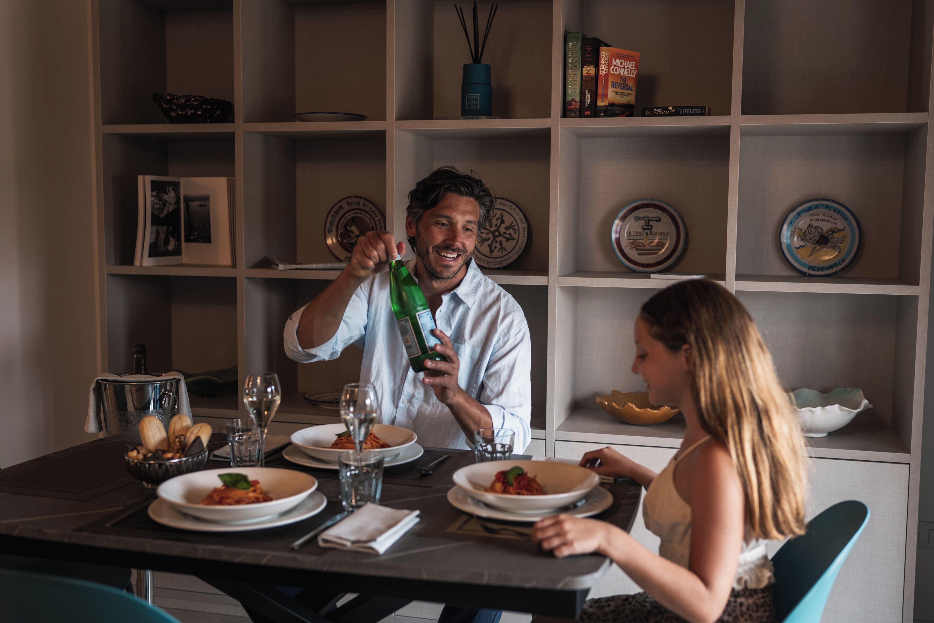 Une table à manger avec des assiettes remplies de nourriture, des verres et une bouteille. On aperçoit, à l'arrière-plan, des étagères avec des assiettes décoratives et un vase.