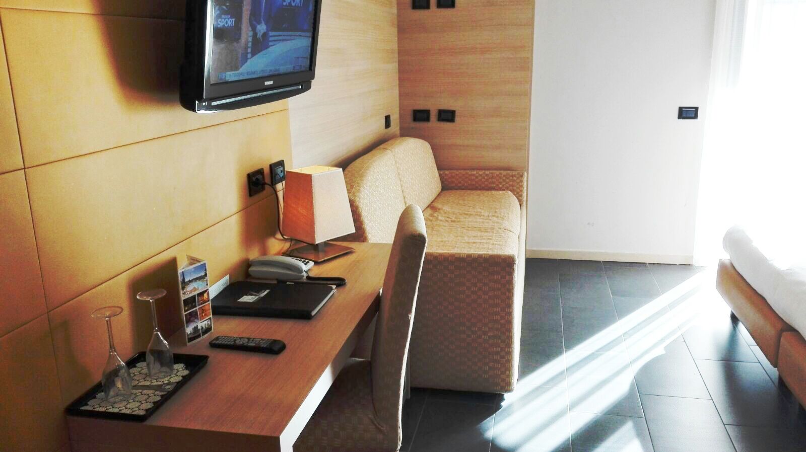 Quarto de hotel moderno com escrivaninha de madeira, TV de tela plana na parede, poltrona confortável, um pequeno sofá e uma janela grande que permite entrada de luz natural.