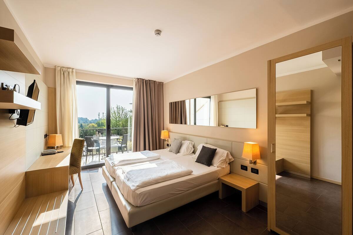 Una habitación de hotel moderna con una cama grande, dos mesitas de noche, un escritorio y una silla. Cuenta con un balcón con vistas a la ciudad.