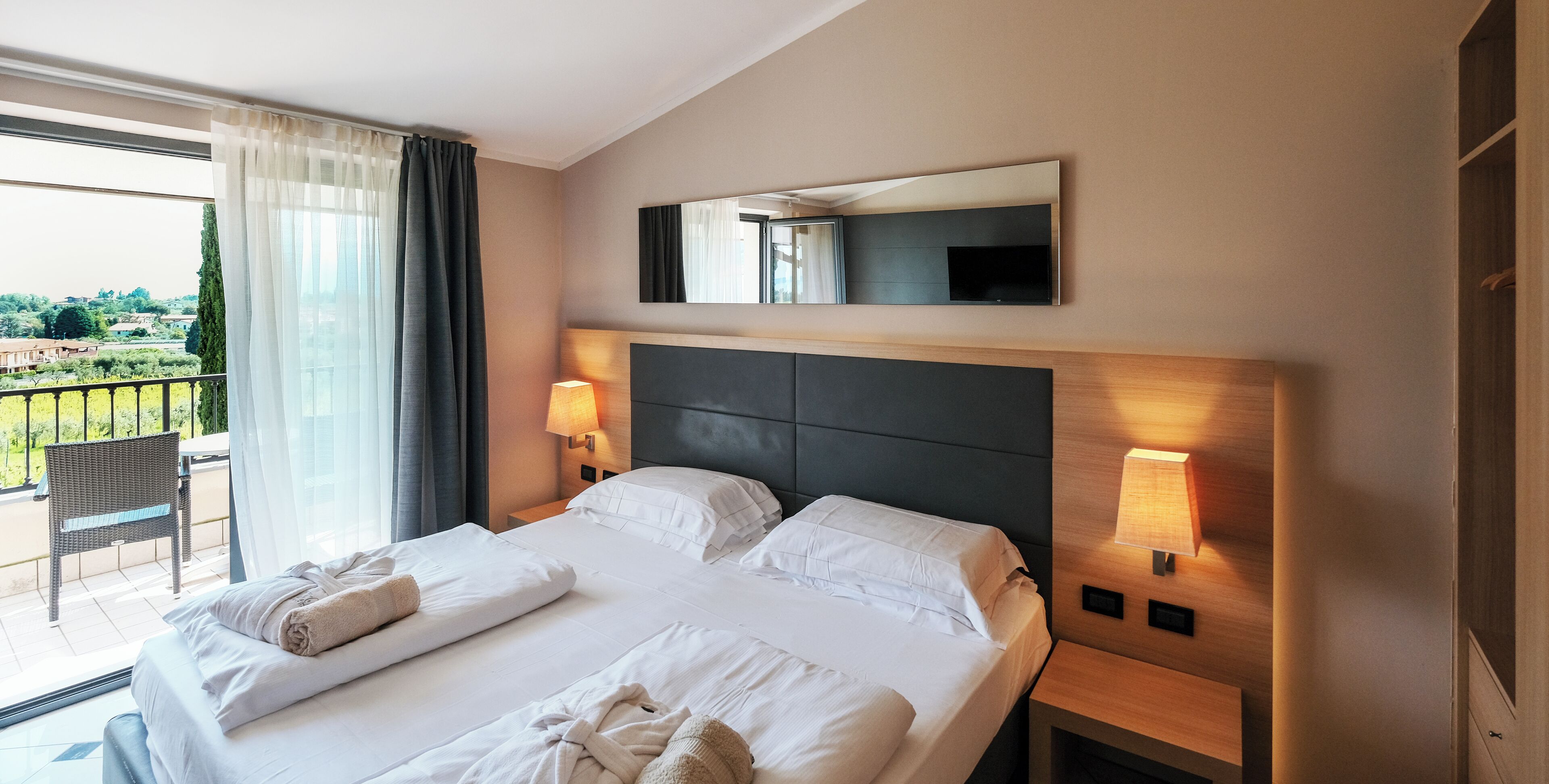 A hotel room with a large bed, two bedside lamps, a mounted TV, and a balcony with a chair.