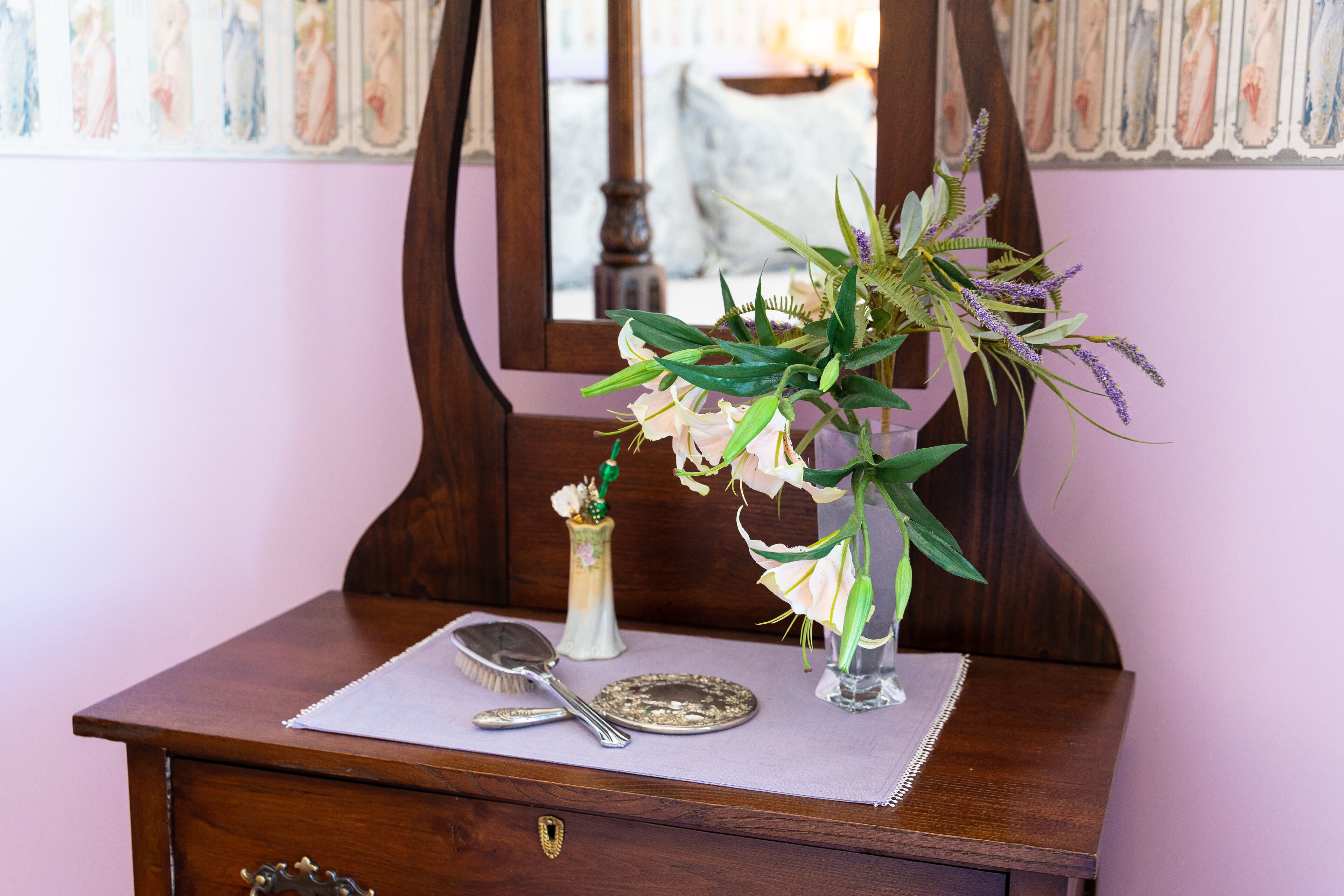 A wooden dresser with a mirror, a vase with flowers, a small vase with a candle, and a plate and spoon on a placemat.