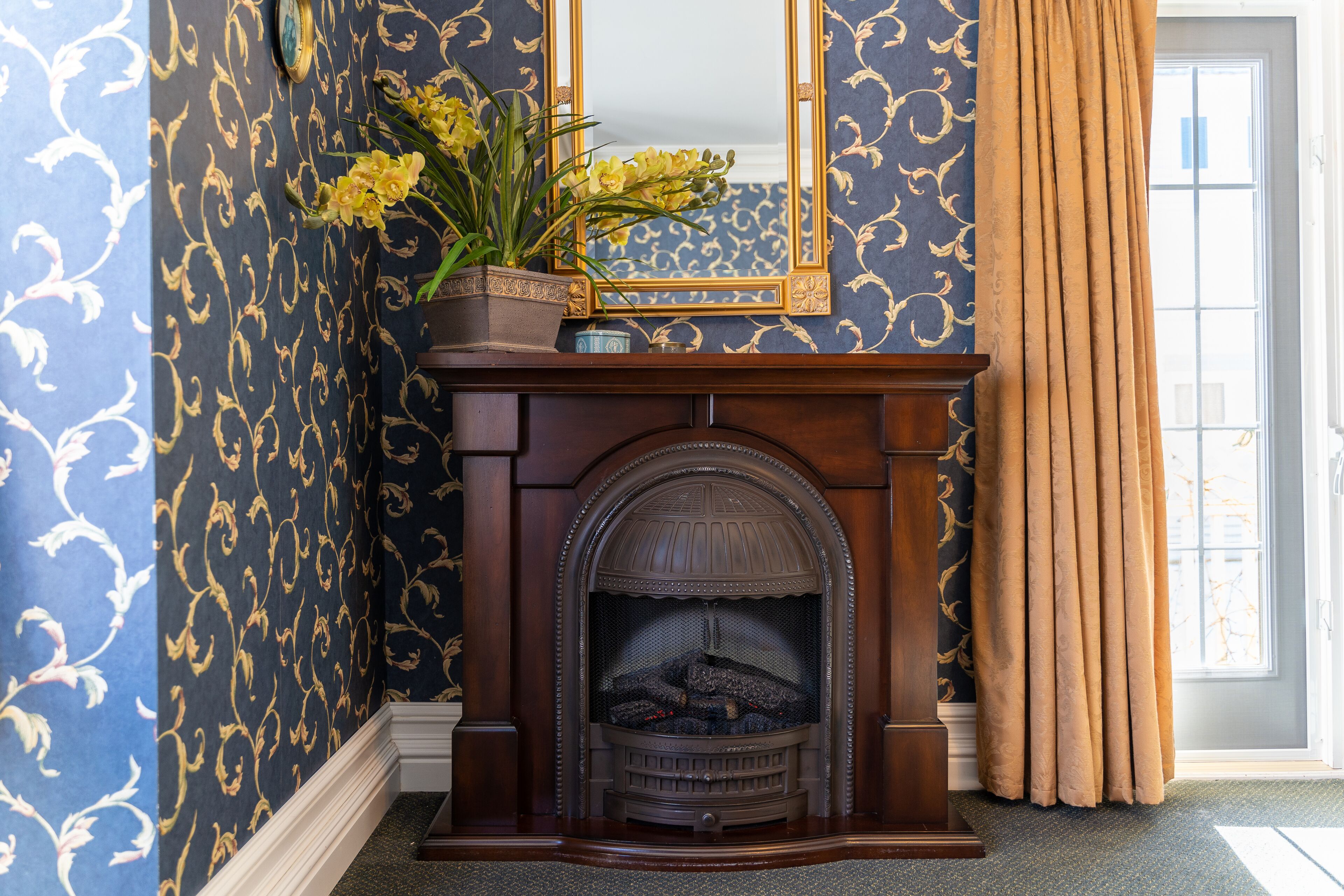 Una habitación con chimenea, papel tapiz floral y un ventanal con cortinas.