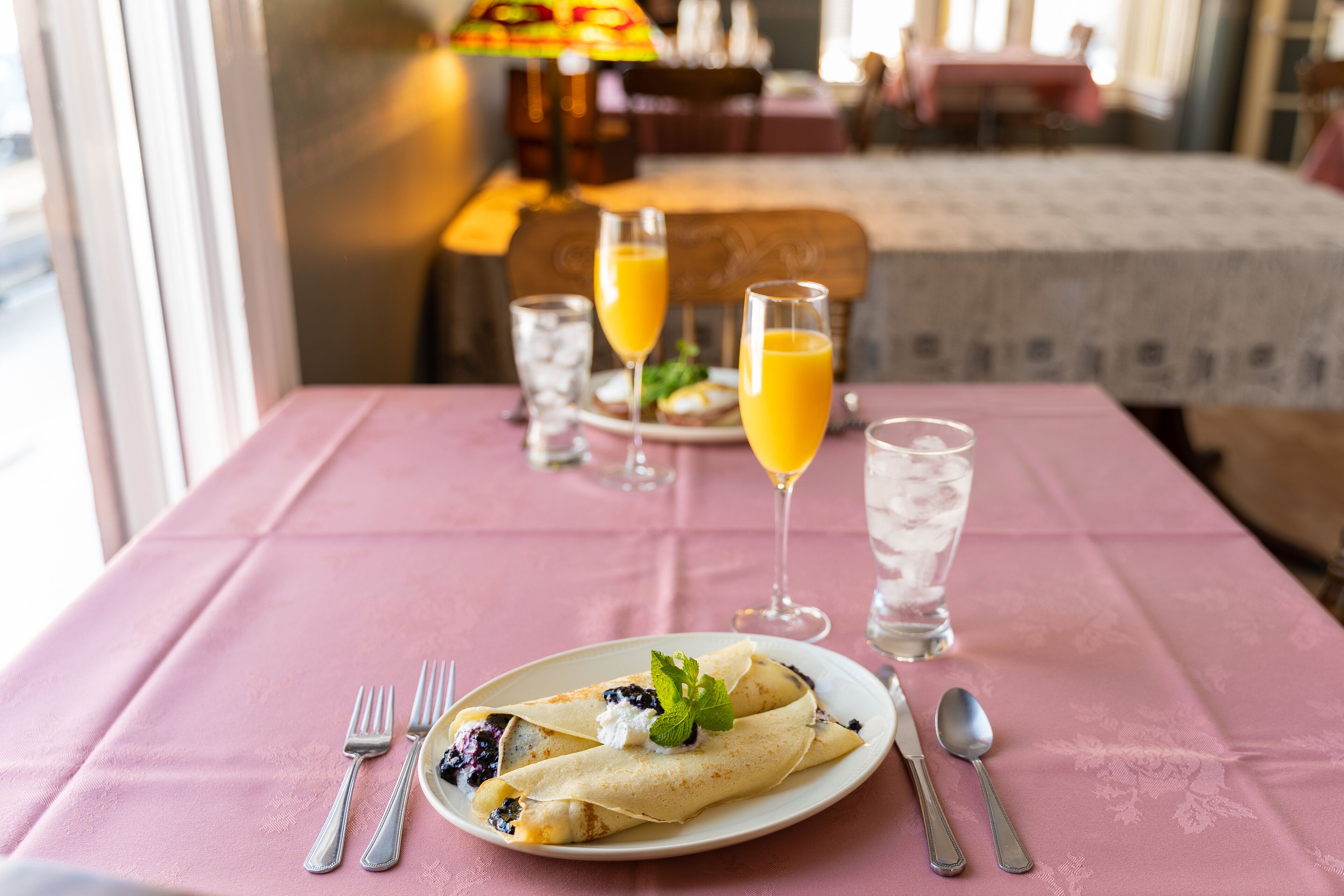 A dining table with a pink tablecloth, a plate of crepes with blueberries and whipped cream, and two glasses of orange juice.