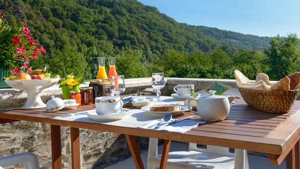 A wooden table set with breakfast items, including fruits, bread, and beverages, overlooking a lush green hillside. - Hostellerie des Lauriers (Curvalle)
