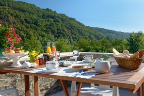 A wooden table set with breakfast items, including fruits, bread, and beverages, overlooking a lush green hillside.