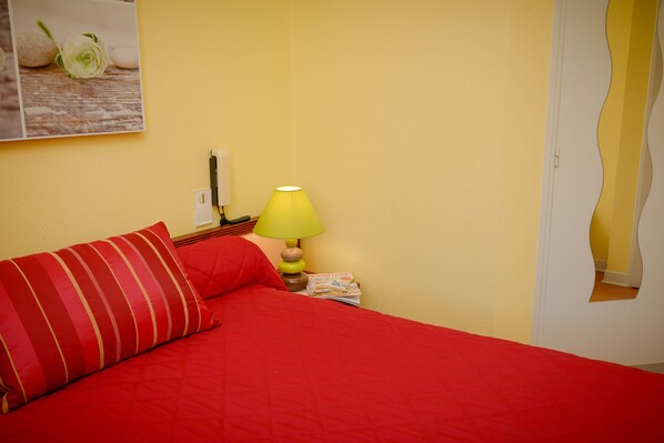 A bed with red bedding, a yellow lamp, and a magazine on the nightstand.