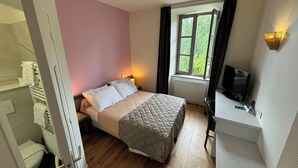 A bedroom with a bed, a desk, a TV, a window with curtains, and a view of greenery outside. - Hostellerie des Lauriers (Curvalle)