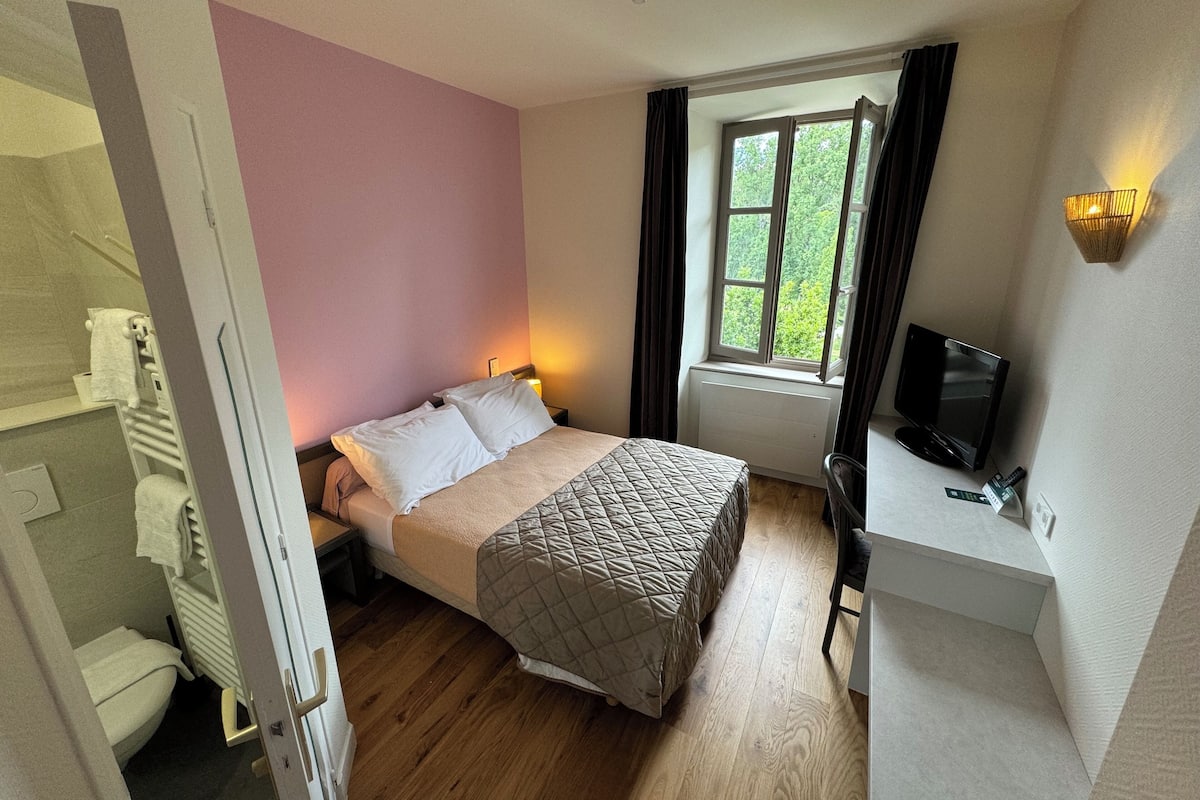 A bedroom with a bed, a desk, a TV, a window with curtains, and a view of greenery outside.
