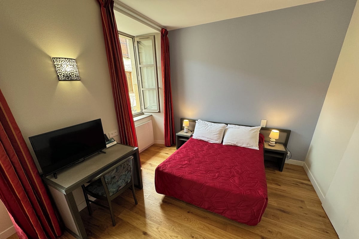 A bedroom with a bed, a desk, a television, and a window with curtains.