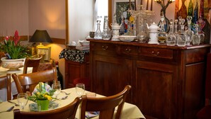 A dining area with a wooden table set for two, surrounded by chairs. A cabinet with glassware and a vase with flowers is visible. - Hostellerie des Lauriers (Curvalle)