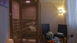 A spa room with a wooden sauna, a massage table, two chairs, a tray with fruits and cups, and a wall-mounted light fixture. - Le Continental (Chateauroux)