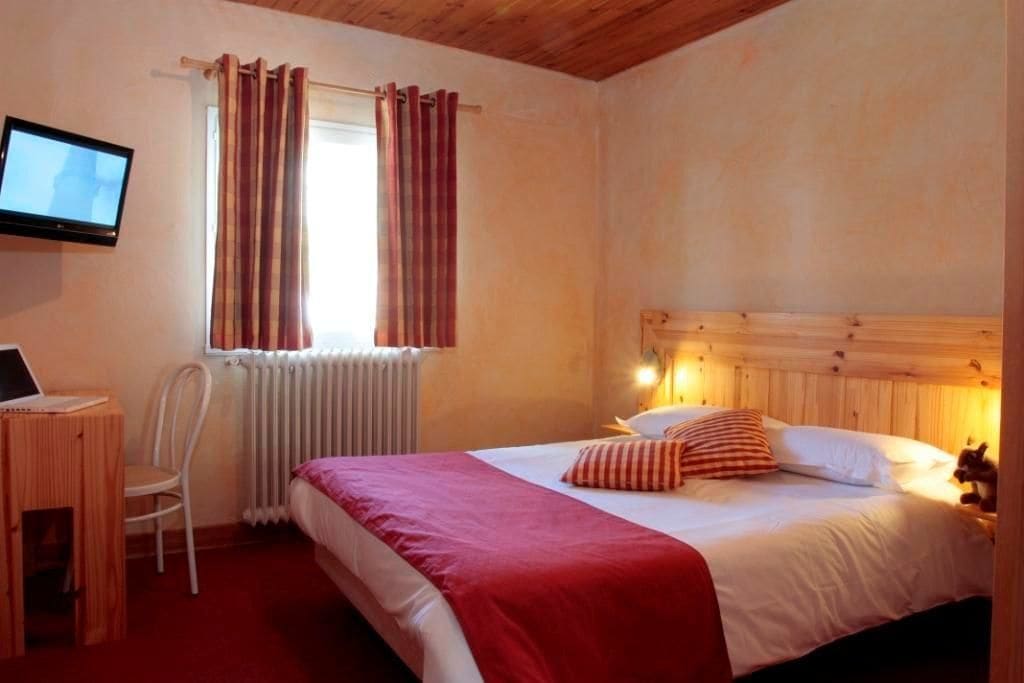 Una camera da letto con un letto, una scrivania con un portatile, una sedia, una finestra con tende e una televisione appesa al muro.
