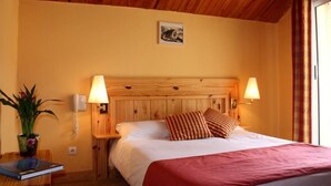 Una habitación de hotel con cabecera de madera, dos lámparas de noche, cama con ropa blanca y una manta roja, una mesita con una planta y un cuadro en la pared.