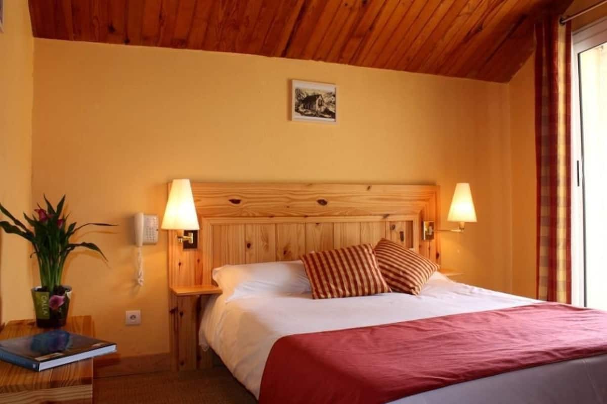 A hotel room with a wooden headboard, two bedside lamps, a bed with white linens and a red blanket, a small table with a plant, and a framed picture on the wall.