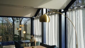 Un comedor moderno con una mesa y sillas de madera, una lámpara de pie dorada y vista a árboles a través de grandes ventanales.