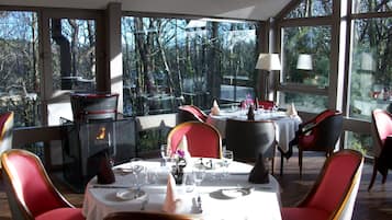 Un comedor con mesas redondas, sillas tapizadas en rojo y vista a árboles a través de amplios ventanales.