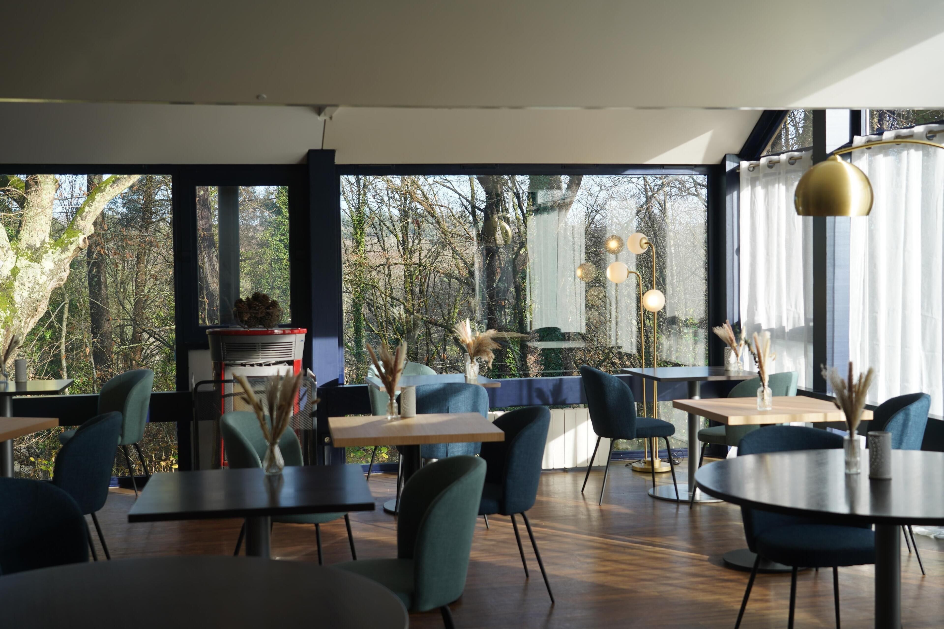 A modern restaurant with a view of trees through large windows. Tables and chairs are arranged for dining.