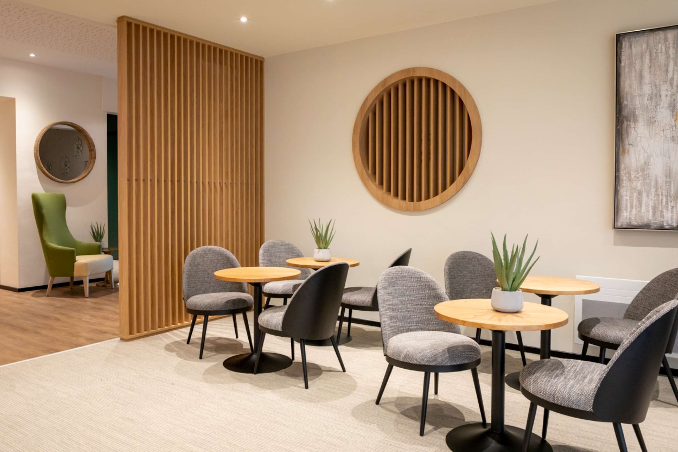 A modern waiting area with round tables, grey upholstered chairs, wooden partitions, and a large abstract painting on the wall.