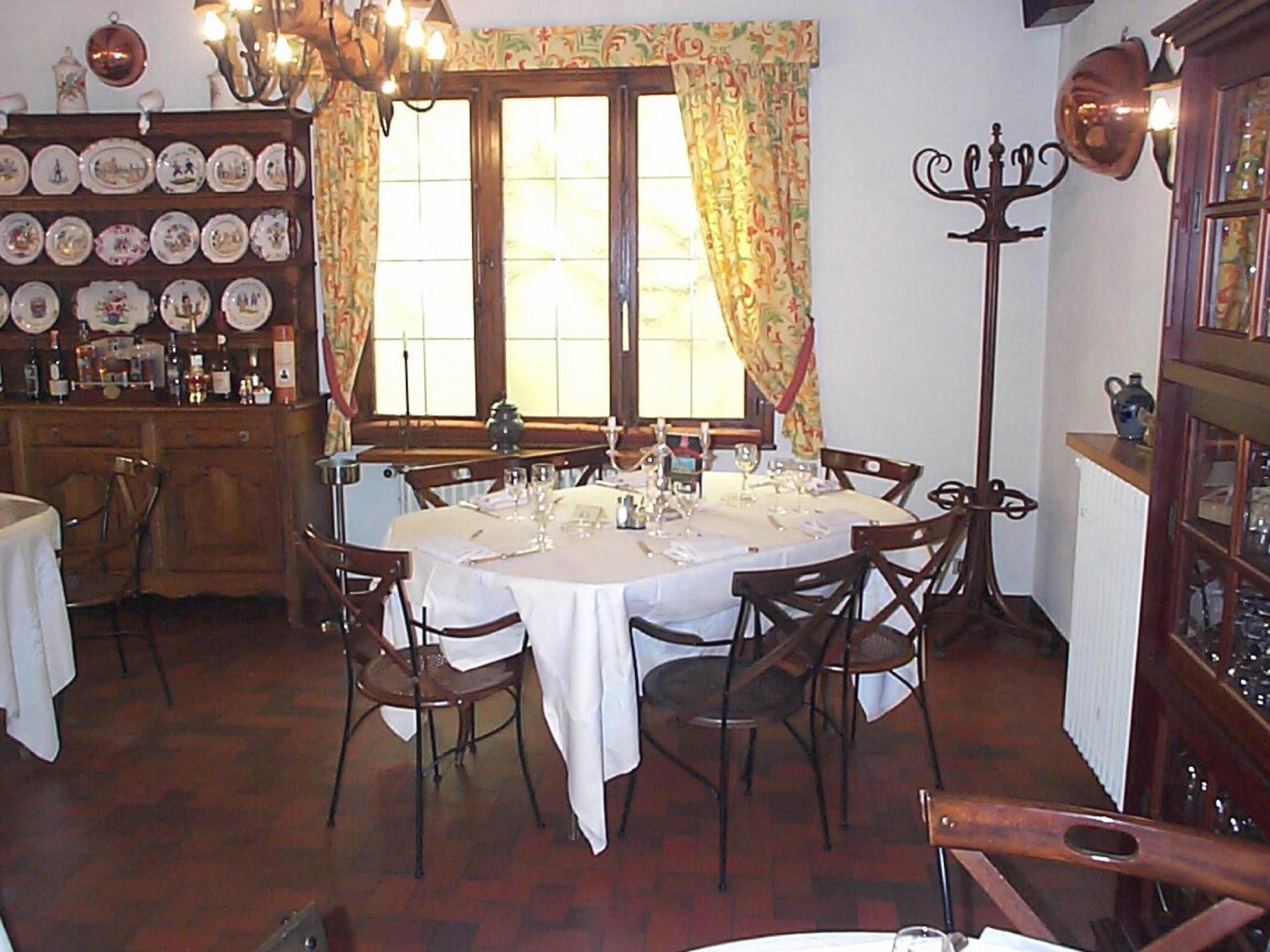 Un coin repas avec une table ronde dressée pour six personnes, entourée de chaises en bois. Un mur décoré d’assiettes orne l’espace au-dessus de la table, et une grande lampe sur pied, richement décorée, se trouve à droite.