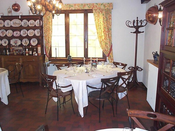 A dining area with a round table set for six, surrounded by wooden chairs. There is a wall of decorative plates above the table, and a tall, ornate floor lamp to the right.