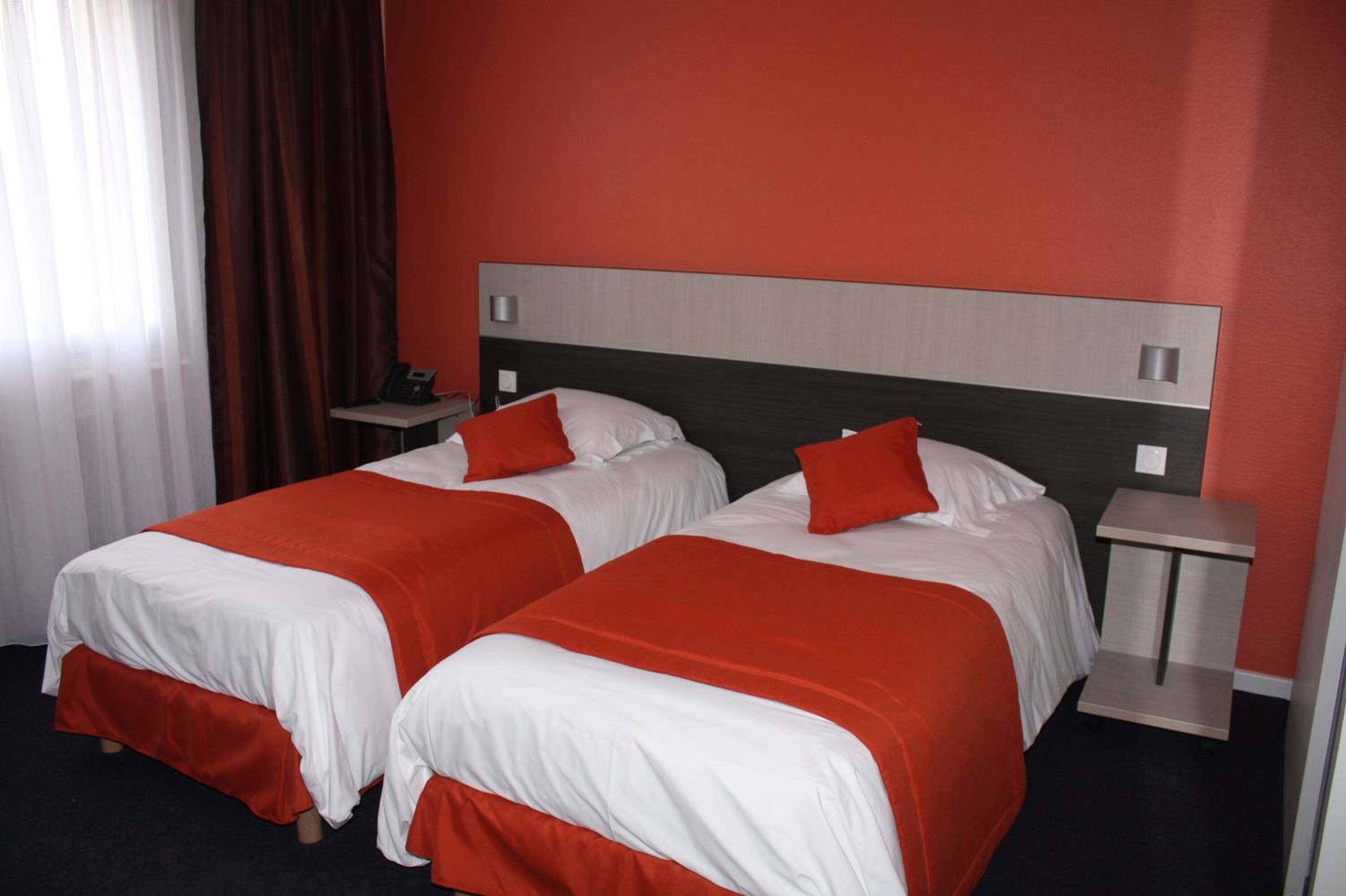 Ein Hotelzimmer mit zwei Betten, jeweils mit weißer und roter Bettwäsche, einem Holzkopfstück und einem kleinen Nachttisch mit Lampe.